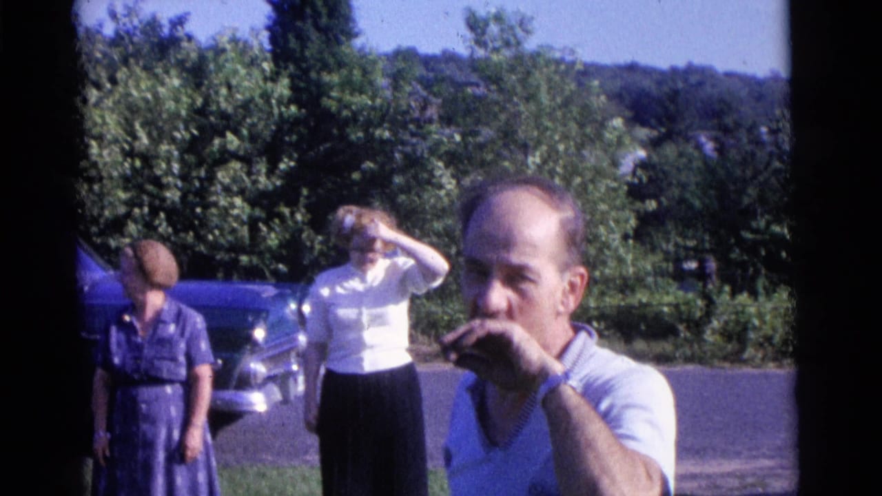 Man near the camera with two women and a parked car behind him outdoors.