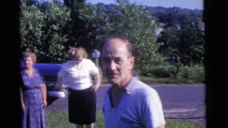1961 Camden home-movie frame showing a man on a grassy lawn, two women nearby, leafy trees, and a parked blue car in the background.