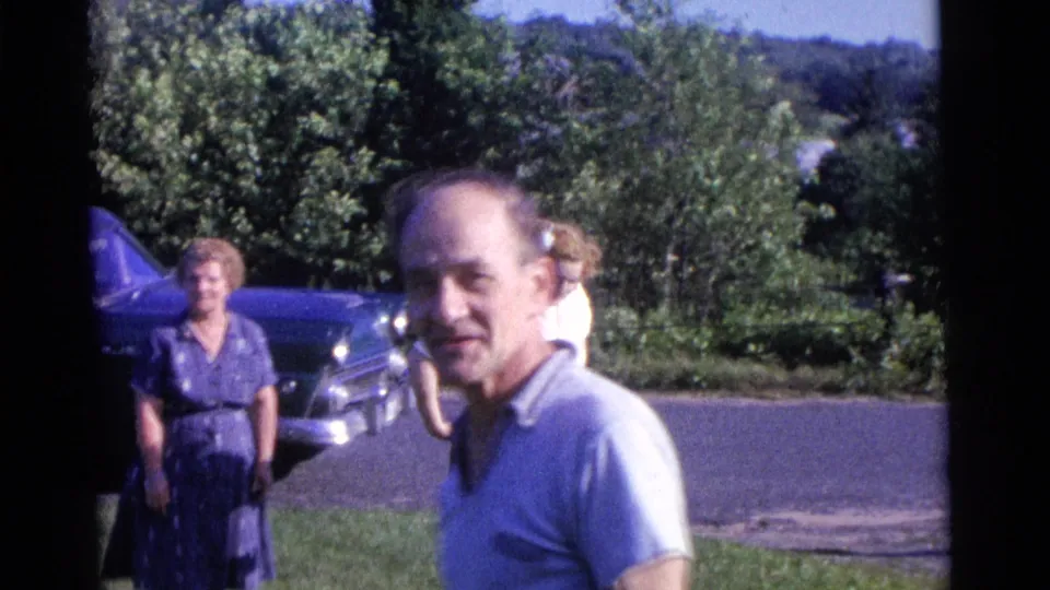 1961 Camden home-movie frame with a man smiling toward the camera, two women in the background, lawn, trees, and a parked blue car.