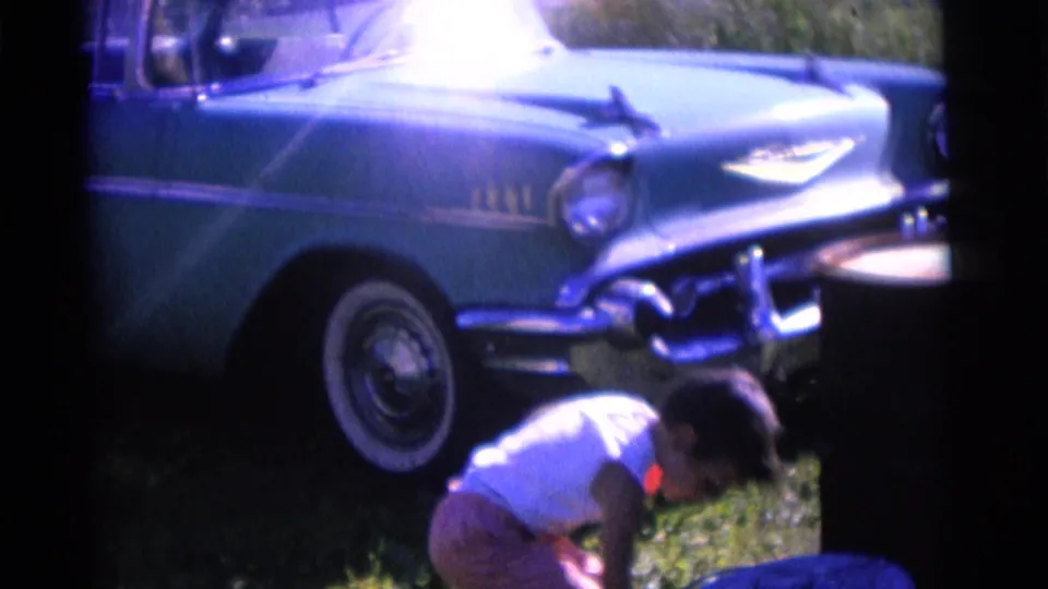 A child crouches in a Camden yard beside a blue Chevrolet Bel Air, with grass and the car's chrome details visible.