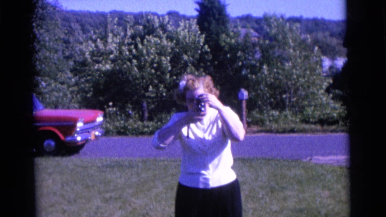 Woman in a white shirt holds a camera to her face on a lawn with a red car behind her.