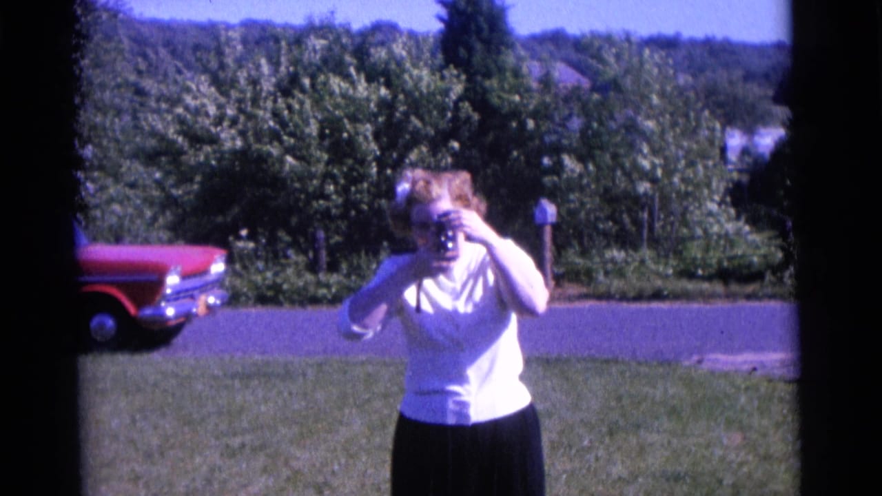 The 1961 Camden home-movie view holds on the woman with camera, framed by a lawn, roadside red car, trees, and distant hillside.