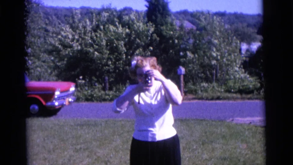 The 1961 Camden home-movie view holds on the woman with camera, framed by a lawn, roadside red car, trees, and distant hillside.
