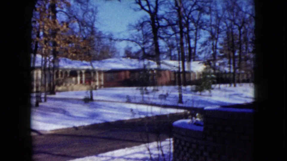 1960 home-movie view of a snow-covered Madison residential street with bare trees, a driveway, and a low house in the background.