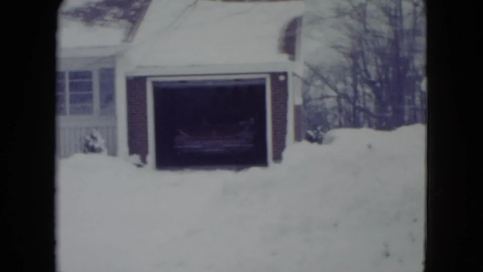 1960 Madison, Wisconsin home-movie frame showing deep snow outside an open brick garage with a car partly visible in shadow.