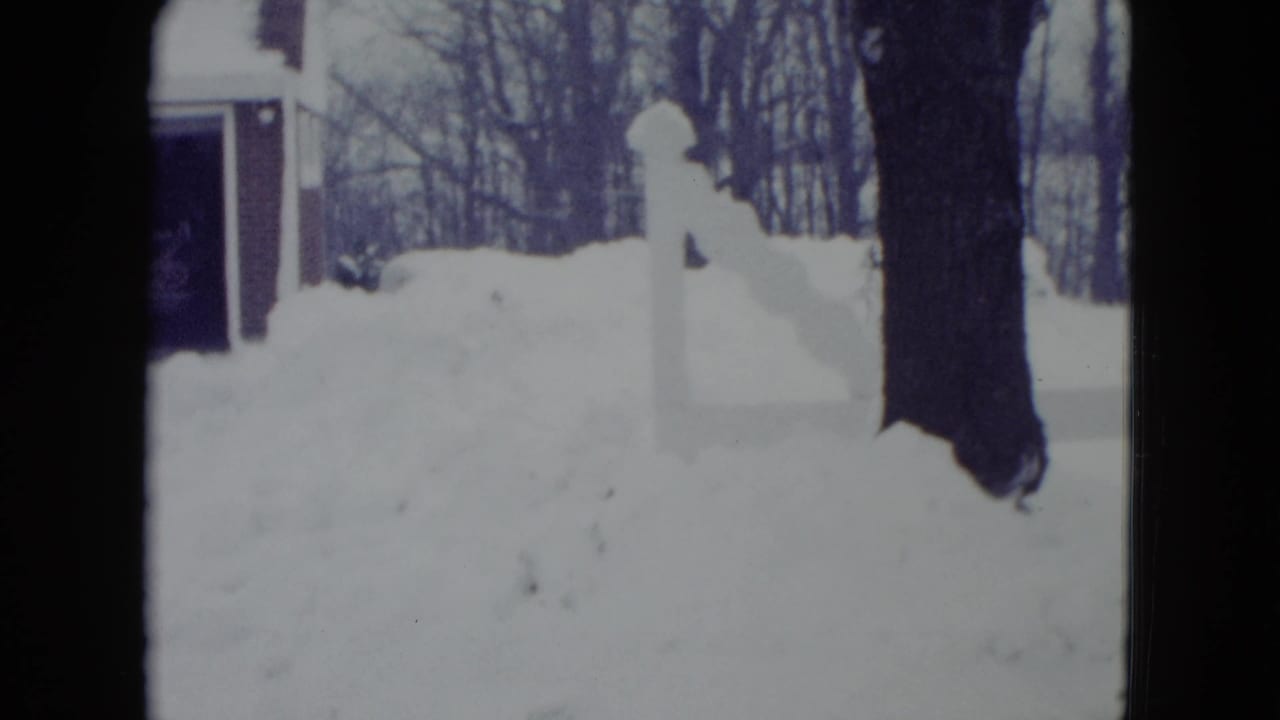 Deep snowbank beside a white fence or post, with bare trees and part of a garage behind it.