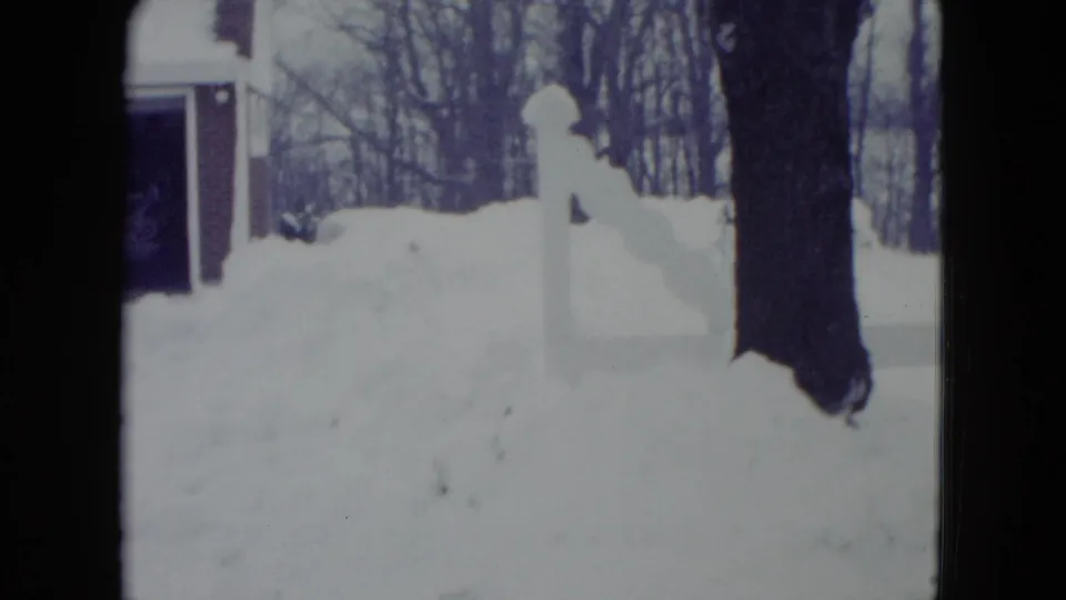 1960 Madison winter yard view with a high snowbank, white fence or post, tree trunk, and part of the garage at the edge of frame.