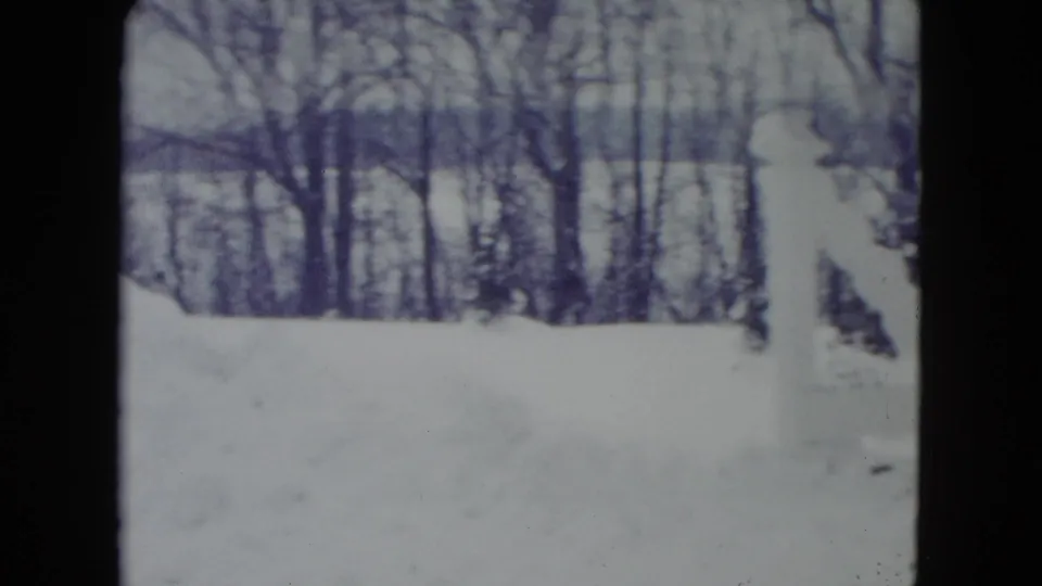 1960 Madison home-movie frame of a snow-covered yard, a white post or fence section, and bare trees beyond the snowbank.
