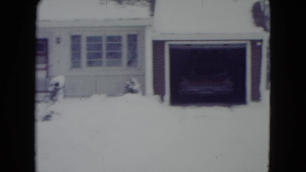 1960 Madison, Wisconsin home-movie frame of a white suburban house with snow, attached garage, front windows, and a dark vehicle inside.