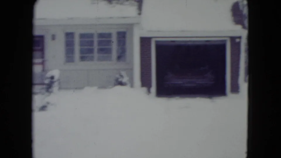 1960 Madison, Wisconsin home-movie frame of a white suburban house with snow, attached garage, front windows, and a dark vehicle inside.