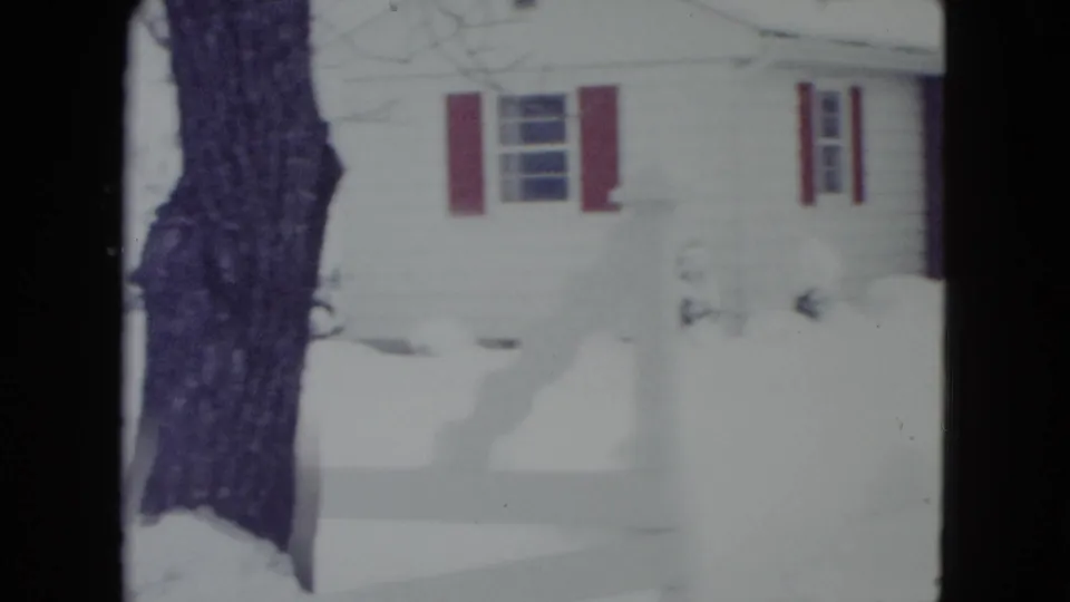 1960 Madison, Wisconsin home-movie frame showing the side of a snowbound white house, red shutters, and a large tree trunk.