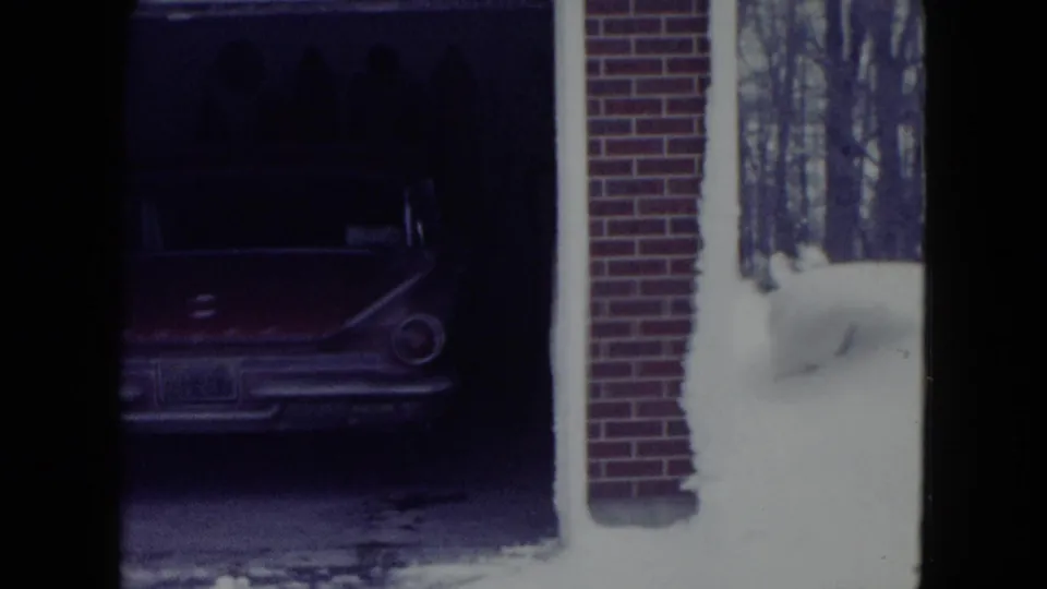 Residential garage view with a parked mid-century car, brick wall, snowdrift, and bare trees in Madison winter footage.
