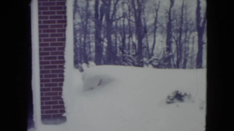 1960 Madison 8mm frame showing a snowy yard, brick building edge, snowbank, and leafless trees in the background.