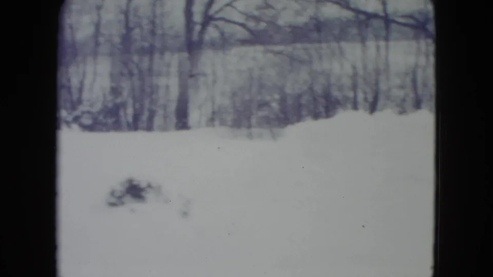 1960 Madison winter frame with deep snow across the yard, bare trees, and a distant wooded hillside.
