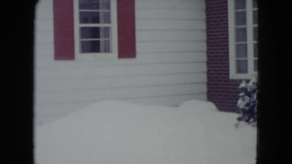Madison 1960 home-movie frame of a snow-covered house exterior with white siding, red shutters, and windows.