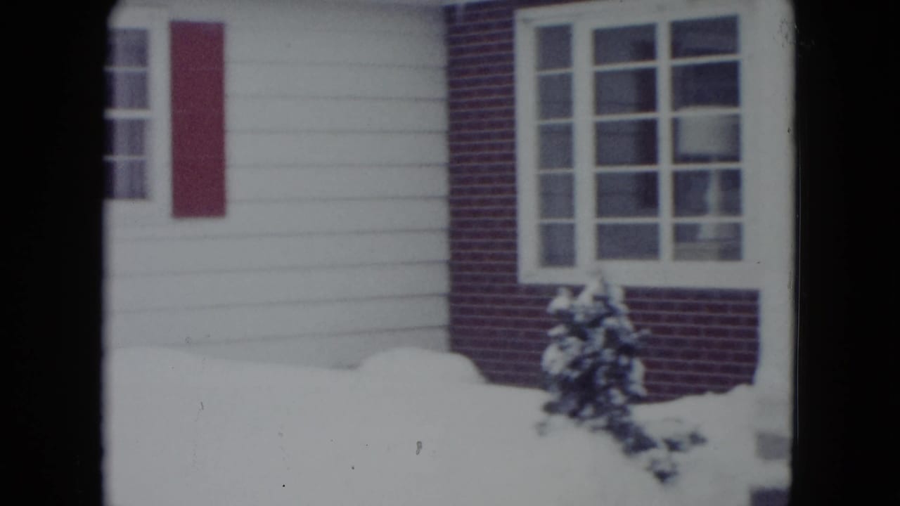Snow-covered house exterior with white siding, red brick, red shutters, windows, and shrubs.