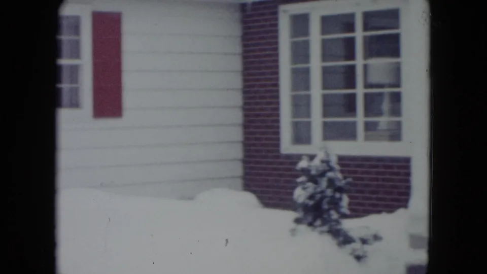 1960 Madison, Wisconsin home-movie frame showing a snowy residential facade with white siding, red brick, red shutters, and evergreen shrubs.