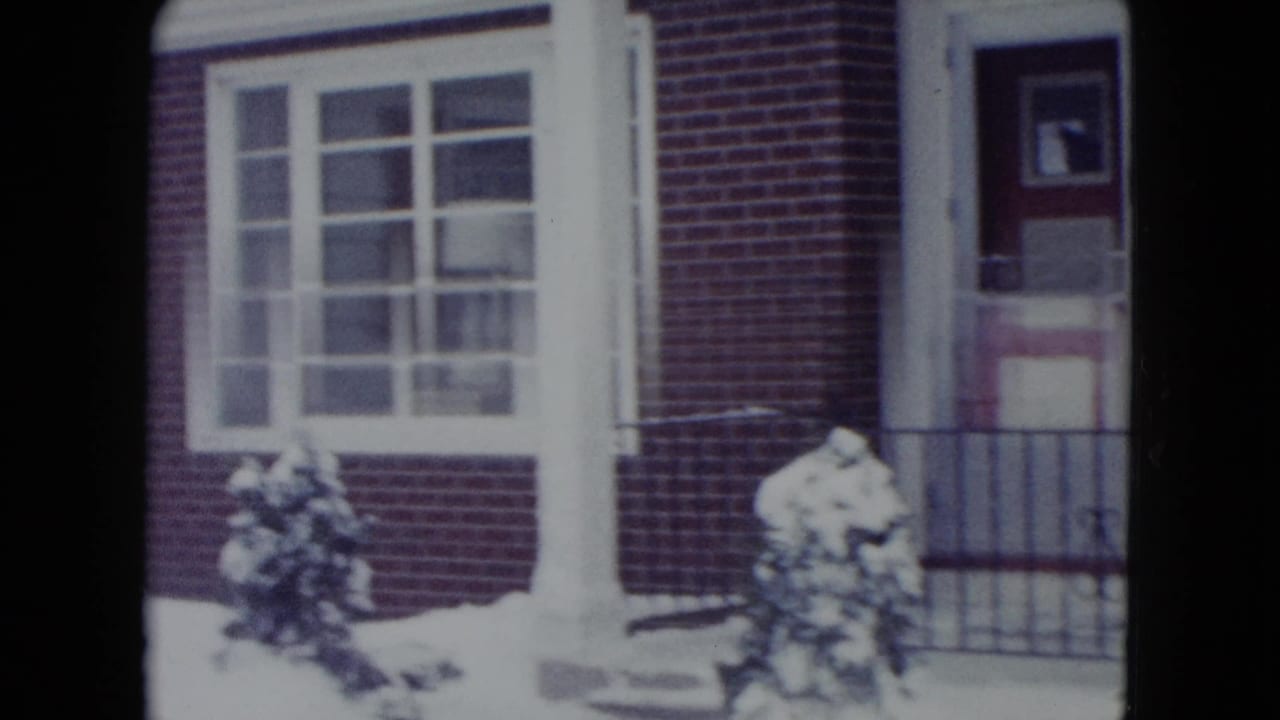 1960 Madison, Wisconsin frame of a snow-covered house entrance with brickwork, multi-pane windows, red door, railing, and shrubs.