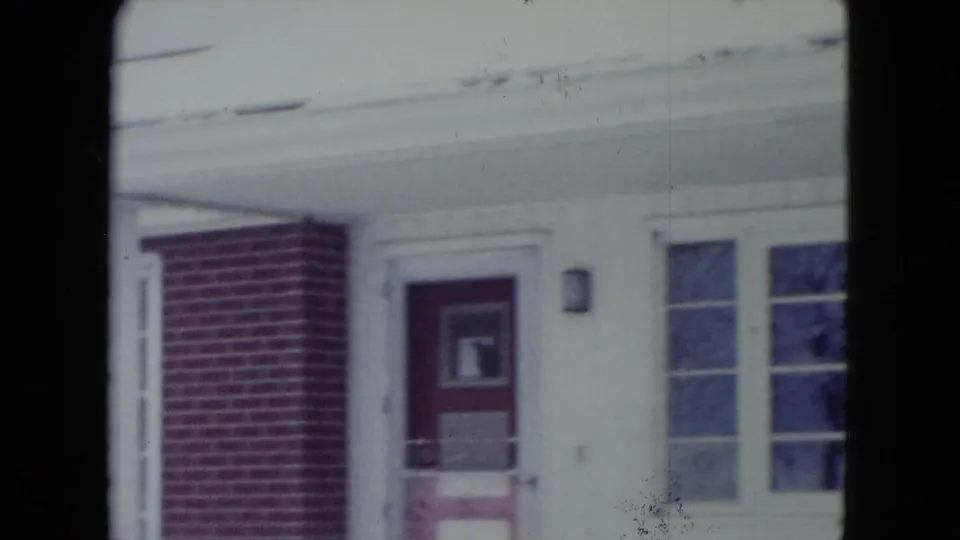 1960 Madison, Wisconsin home-movie frame showing a snowy front porch, red door, brick column, white trim, and windows.