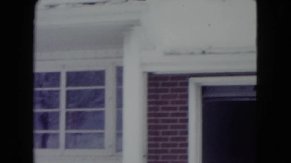 1960 Madison, Wisconsin frame showing the house facade during snow, with a white porch column, large windows, brickwork, and deep shadow at right.