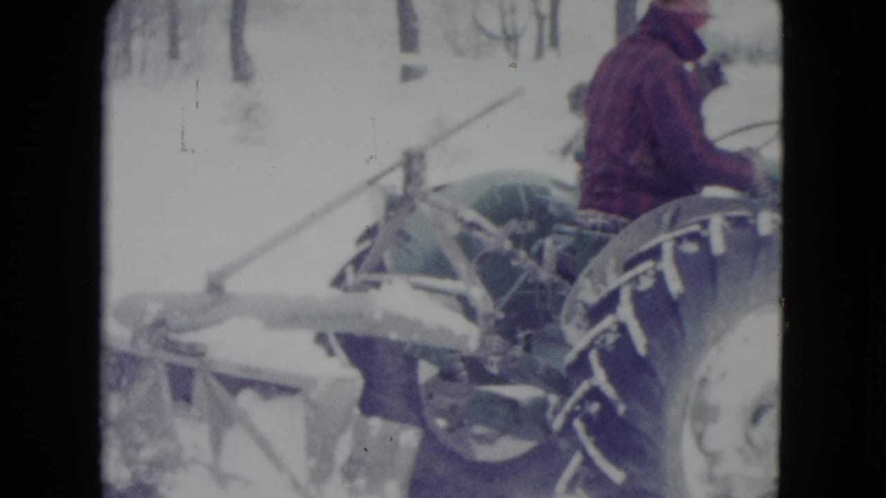 Man in a red jacket sits on a green tractor in deep snow.
