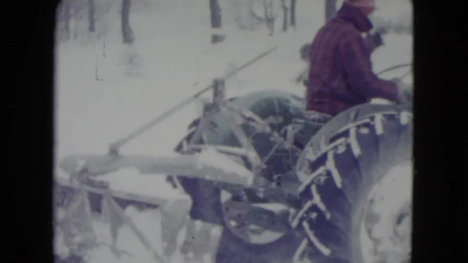 Man operating a green tractor in a snowy Madison, Wisconsin setting, with a rear blade or implement partly visible.