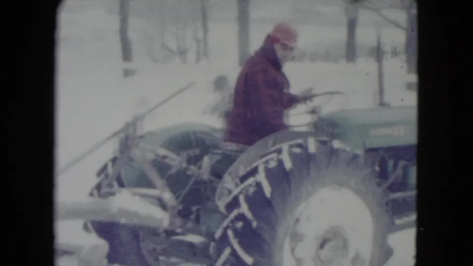 1960 home-movie frame of a man driving a green tractor through deep snow near Madison, with winter trees behind him.