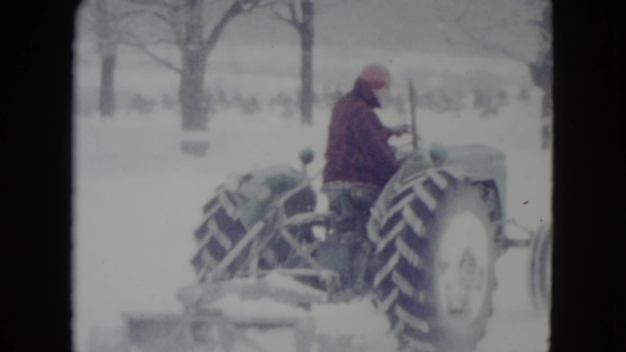 Man in a red jacket drives a tractor through deep snow near bare trees.