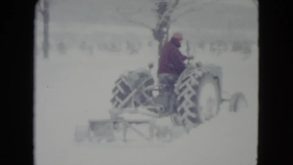 The tractor continues across the snow in rural Madison, Wisconsin, with the driver seated above large rear tires and a plow trailing behind.