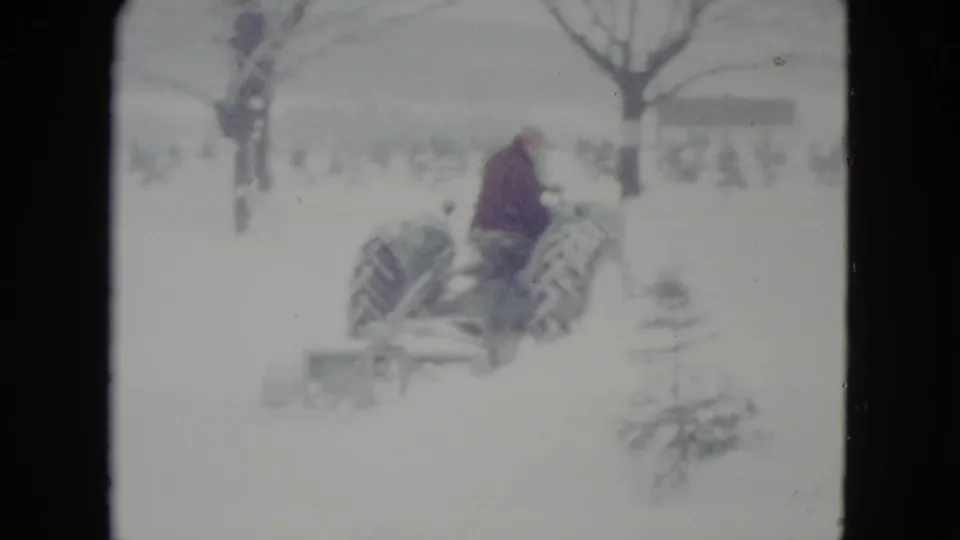 A wider 1960 home-movie view shows the tractor farther away, moving through snow near leafless trees and small evergreens.
