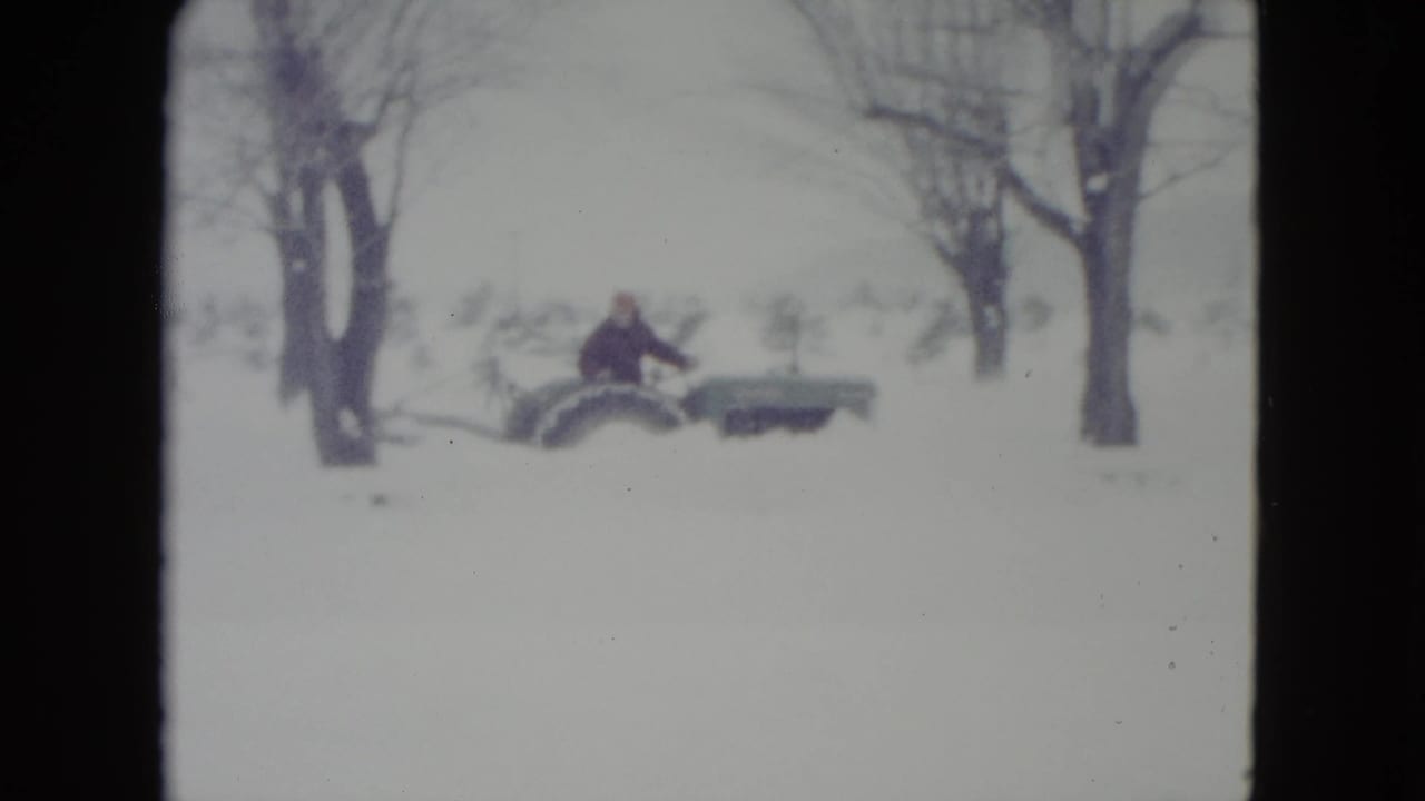 Person on a tractor crosses a snow-covered field with trees behind.
