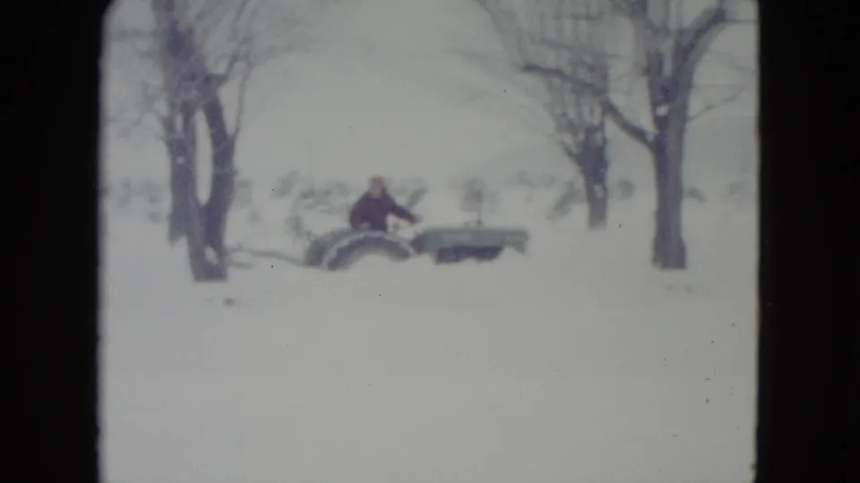 A distant adult in a dark coat and red cap drives a tractor across snowy rural ground in Madison, Wisconsin archival footage.