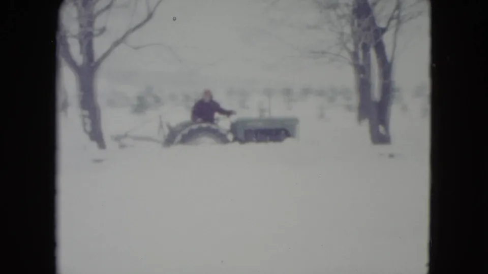 A vintage tractor with a bundled adult operator moves through deep snow between leafless trees in 1960 Madison, Wisconsin footage.