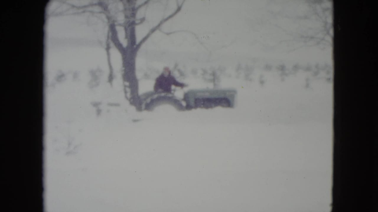 1960 Madison, Wisconsin 8mm frame showing a man near a green tractor in deep snow beside bare trees.