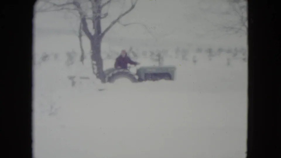 1960 Madison, Wisconsin 8mm frame showing a man near a green tractor in deep snow beside bare trees.