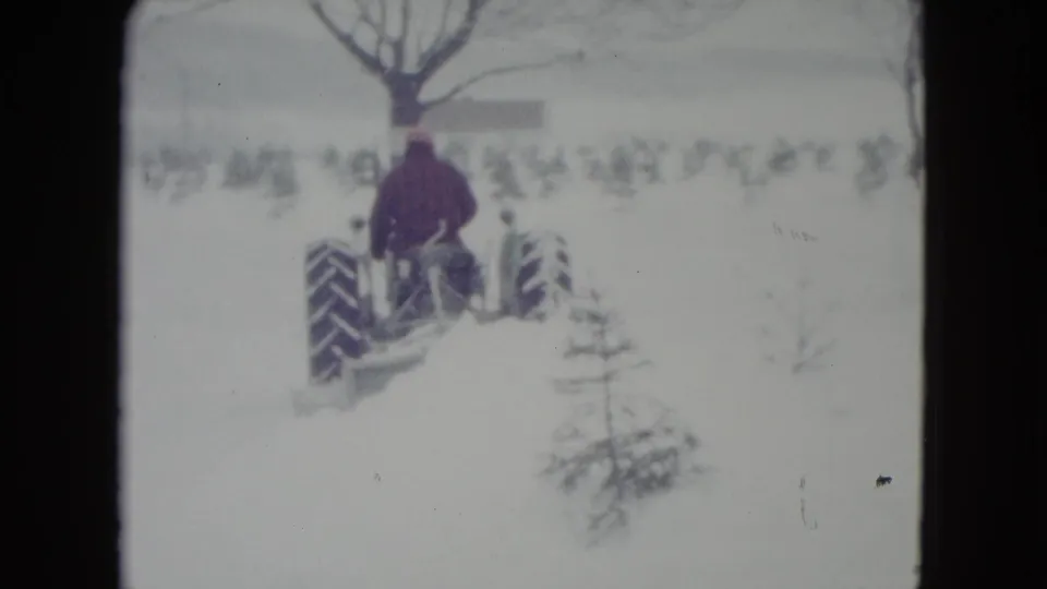 1960 Madison, Wisconsin 8mm frame showing a man driving a tractor through deep snow toward a bare tree.
