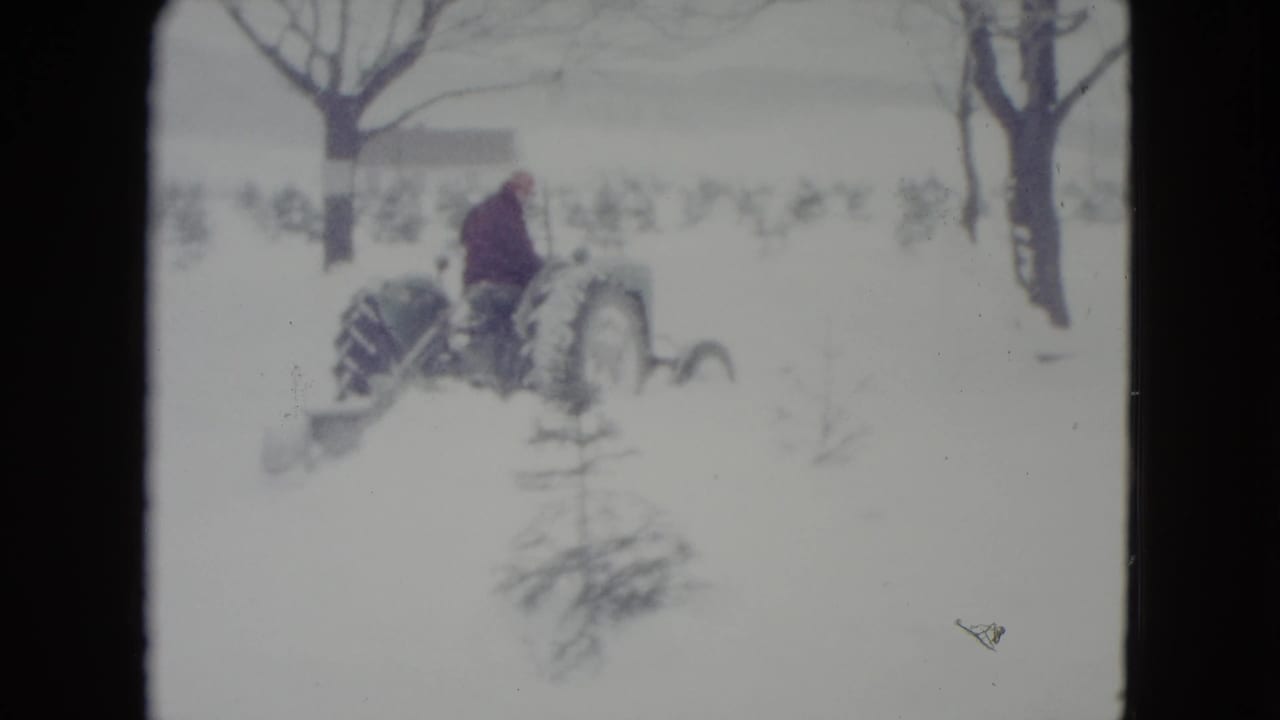 Tractor and driver pass through a snowy field with leafless trees behind them.