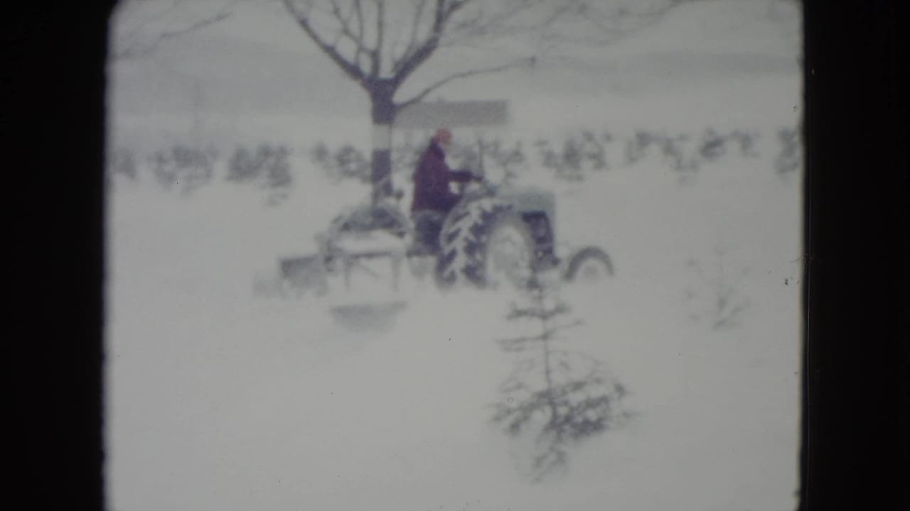 In Madison, Wisconsin, 1960, a bundled adult operates a small tractor with a blade implement across a snowy field.
