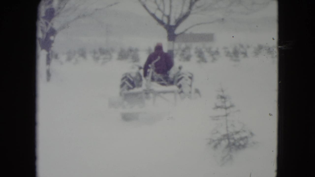 Tractor seen from behind as it moves through a snowy field with trees in the background.
