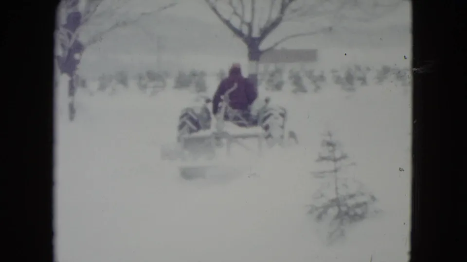 The tractor continues across the Madison snowfield, with its rear wheels and blade visible against bare trees and small evergreens.