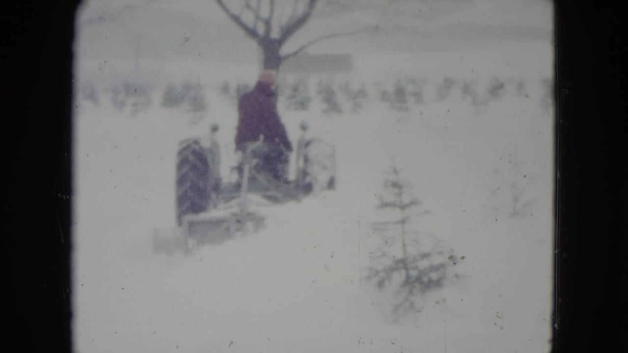 In Madison, Wisconsin, circa 1960, a man in dark clothing rides a vintage tractor through deep snow near bare winter trees.