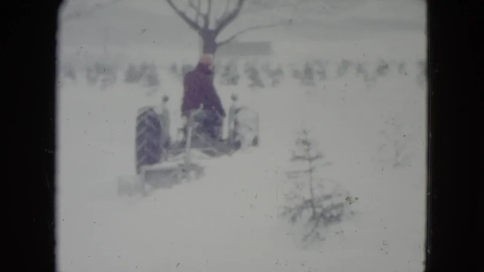 In Madison, Wisconsin, circa 1960, a man in dark clothing rides a vintage tractor through deep snow near bare winter trees.