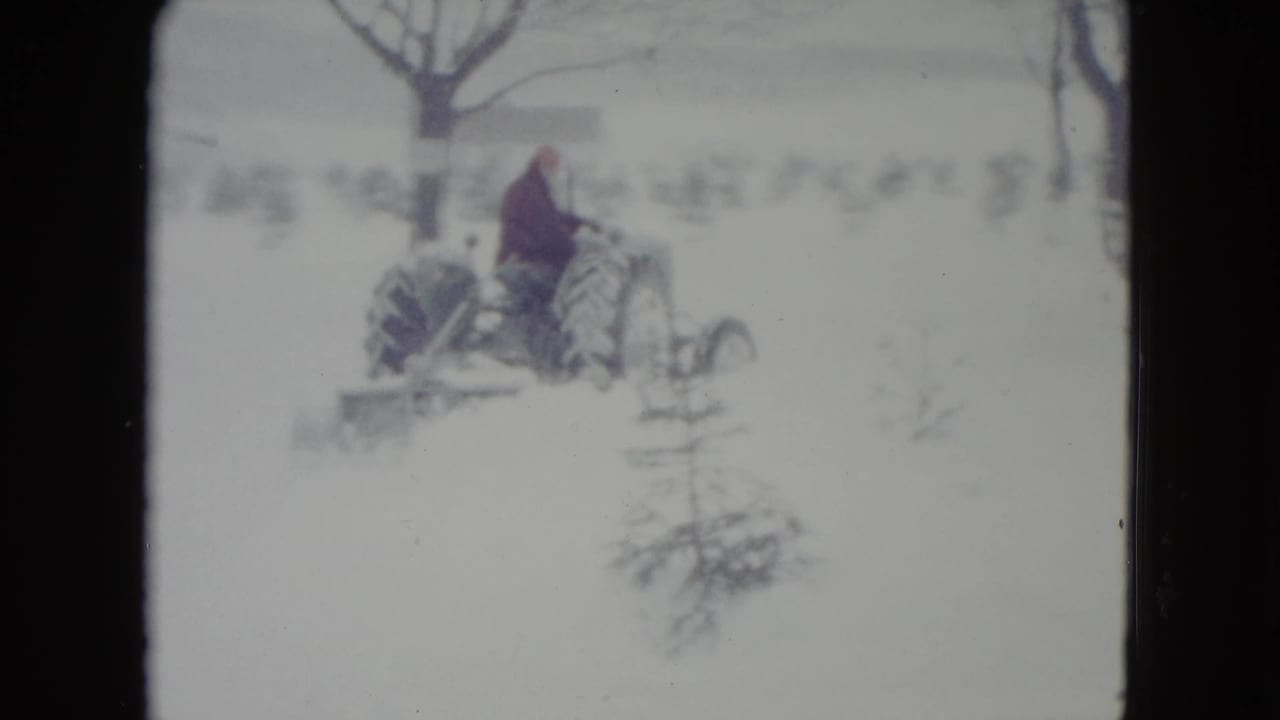 Side view of a tractor crossing snow with a man seated at the controls.