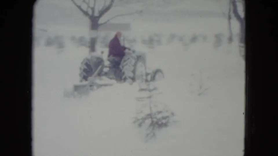 A man rides a vintage tractor across a snowy Madison, Wisconsin field, with bare trees and small evergreens in the background.
