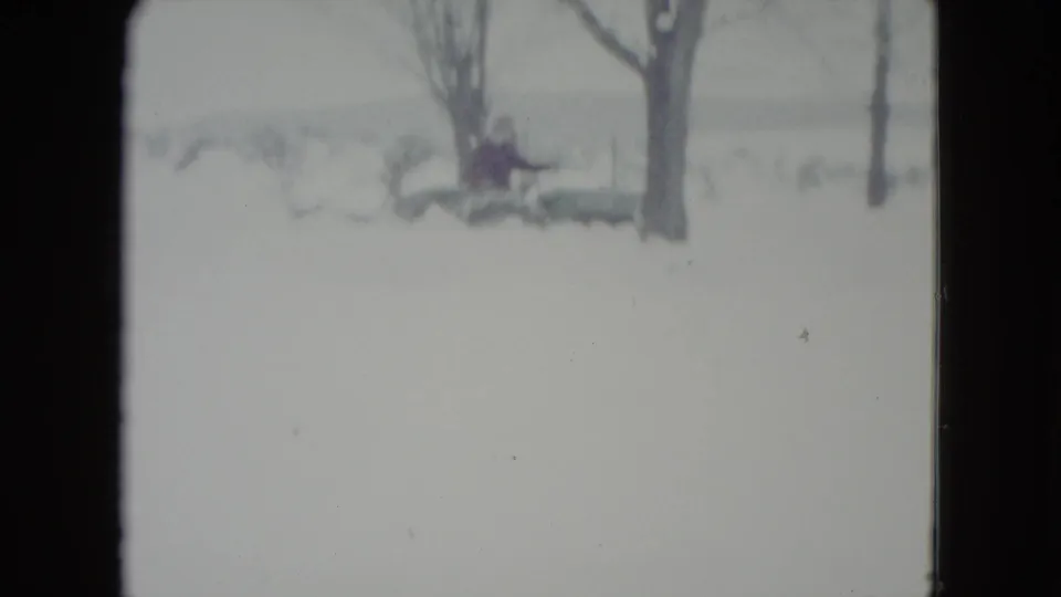 Distant 1960 Madison, Wisconsin winter frame with a person in dark clothing operating a tractor or plow vehicle in deep snow.