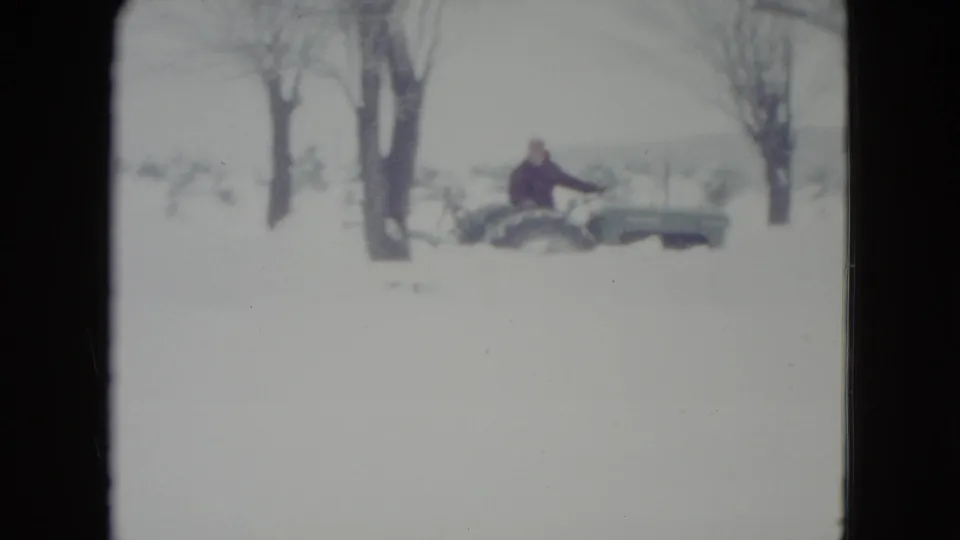 1960 Madison, Wisconsin 8mm frame showing a distant person driving a green tractor or plow vehicle past bare trees in deep snow.