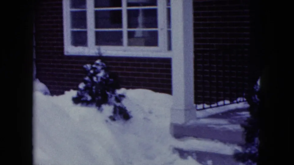 1960 Madison, Wisconsin 8mm frame showing a snow-covered residential exterior with brick wall, window, porch post, and a small shrub.