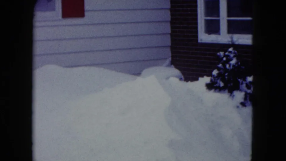 1960 Madison, Wisconsin 8mm frame of a residential exterior with siding, brick, windows, shrubs, and piled snow near the entrance.