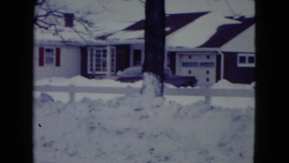 A dark dog is seen low in the snow near a Madison residence, with a garage door, tree trunk, fence, and snowbanks in view.