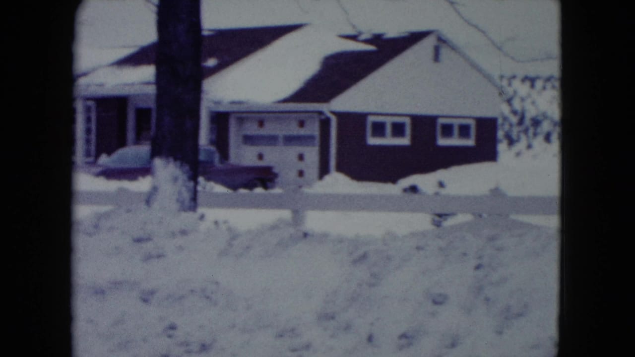 1960 Madison, Wisconsin home-movie frame showing a snow-covered suburban house, driveway car, white fence, and bare winter trees.